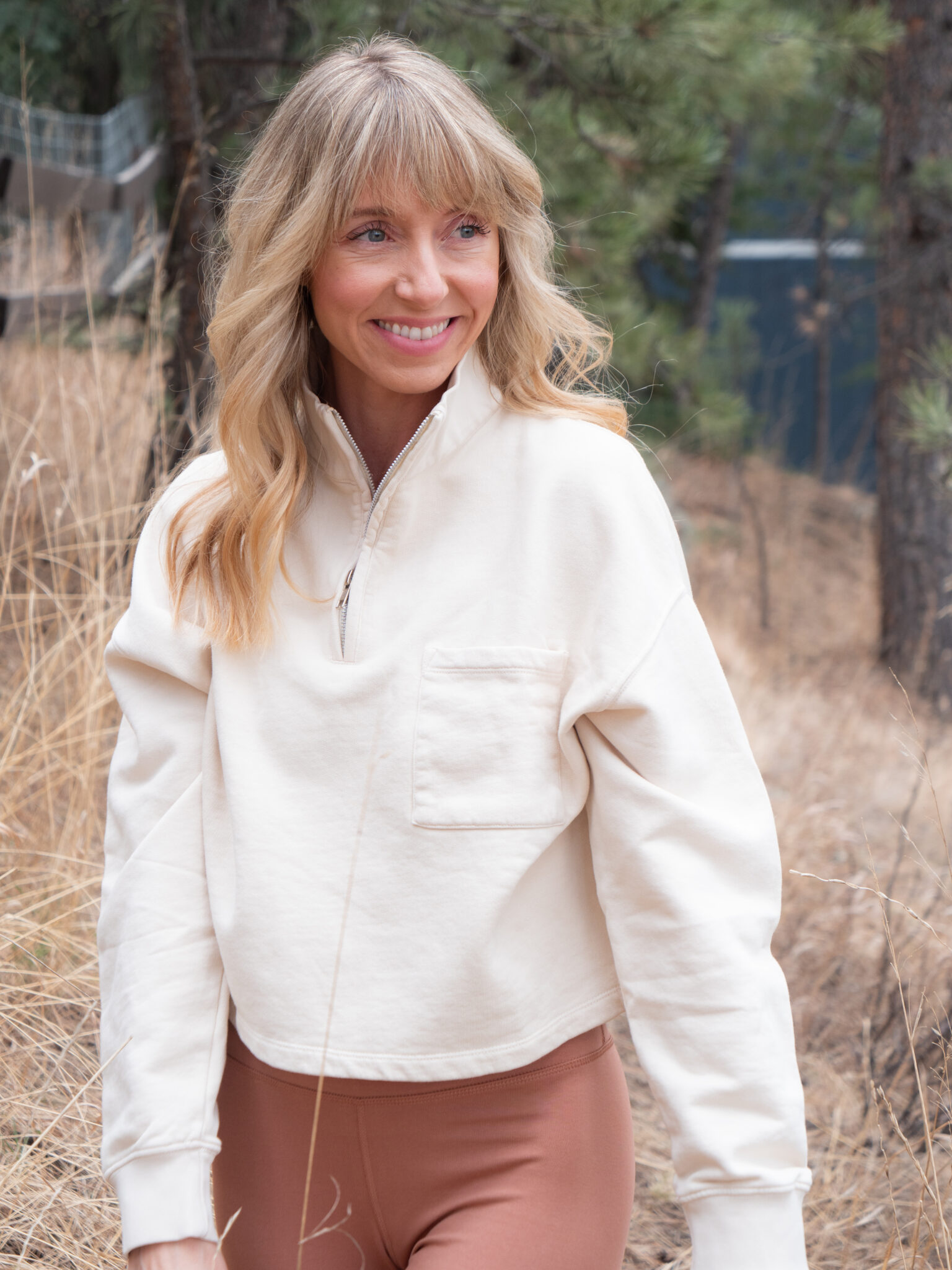 A woman with blonde hair wearing a cream pullover and brown leggings stands outdoors among dry grass and trees, smiling and looking to the side.