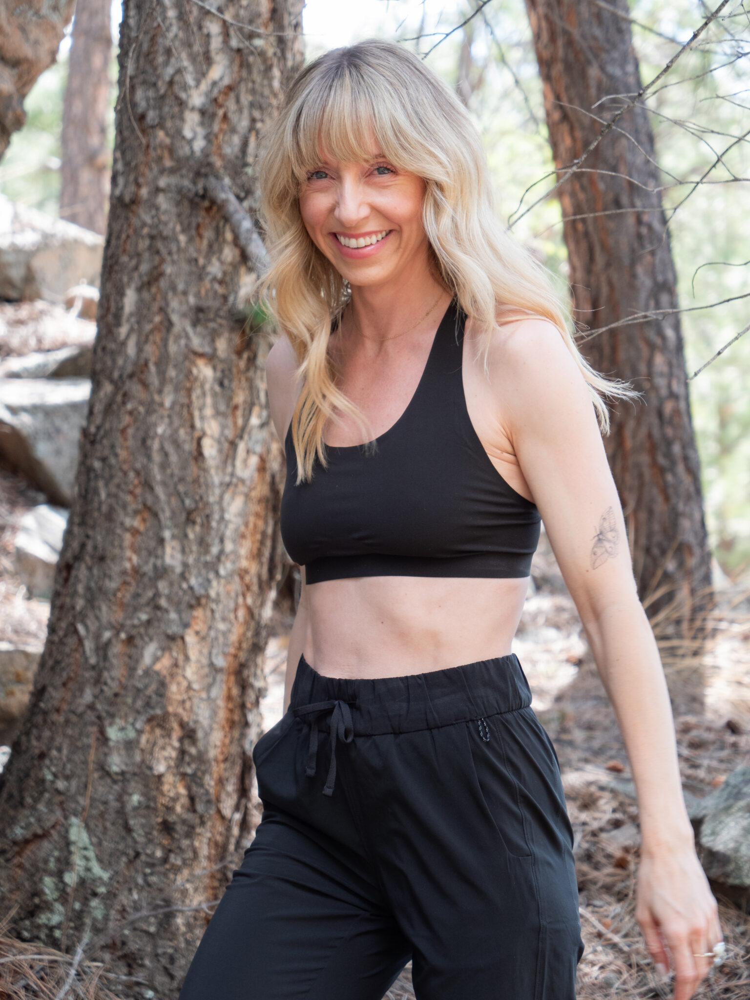 A woman with long blonde hair, wearing a black sports bra and black pants, stands outdoors in a wooded area, smiling at the camera.