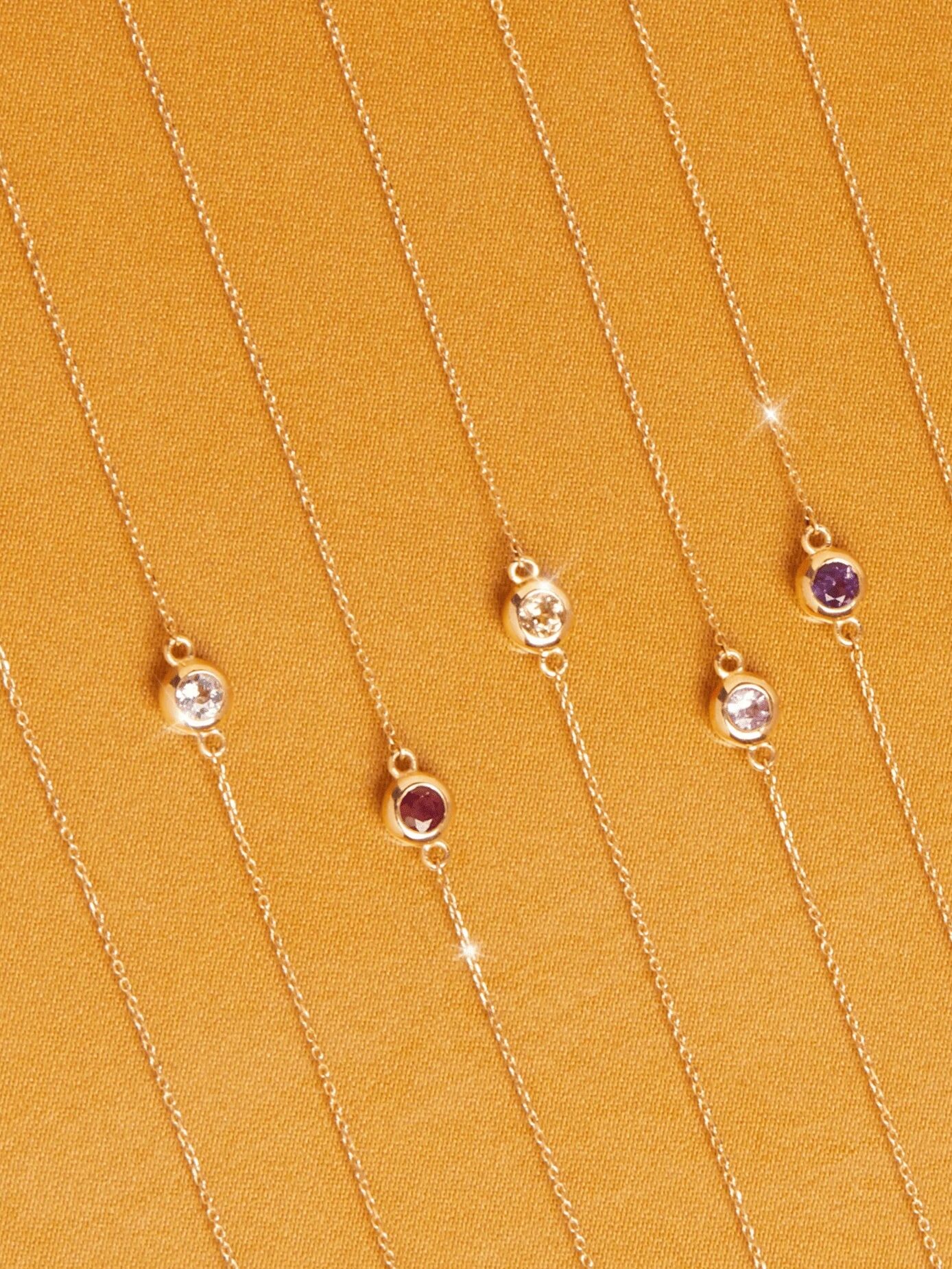 Six gold chain necklaces with round gemstone pendants arranged in parallel lines on a textured golden surface.