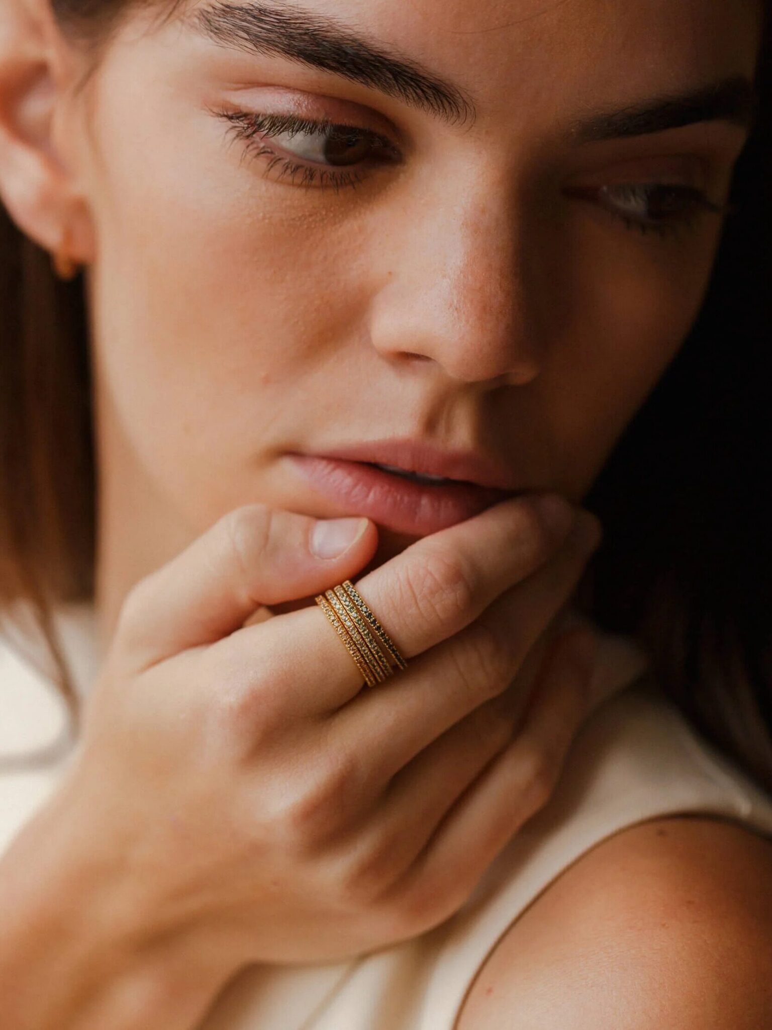 A woman with long brown hair rests her chin on her hand, showing several stacked gold rings on her fingers.