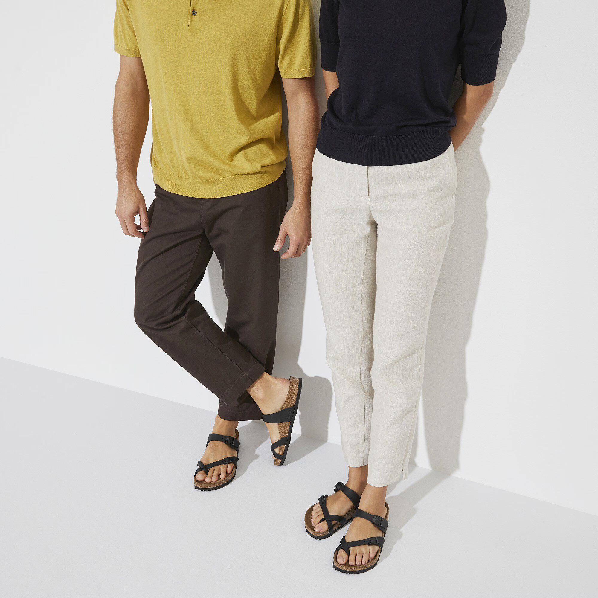 Two people standing side by side, both wearing sandals; one in a yellow shirt and brown pants, the other in a black shirt and light pants against a plain background.