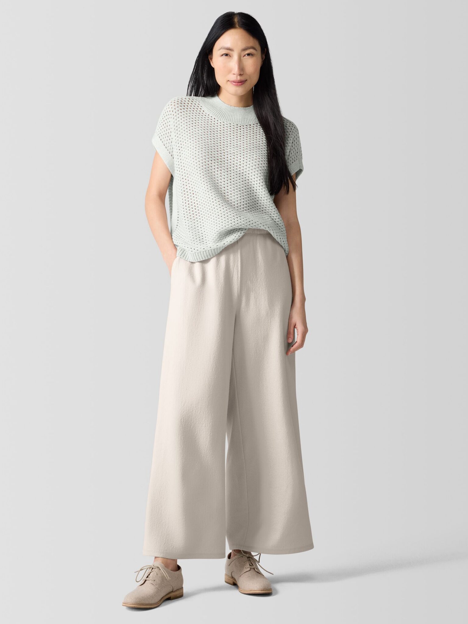 A woman stands against a plain background wearing a light knitted short-sleeve top, wide-leg beige pants, and beige lace-up shoes.
