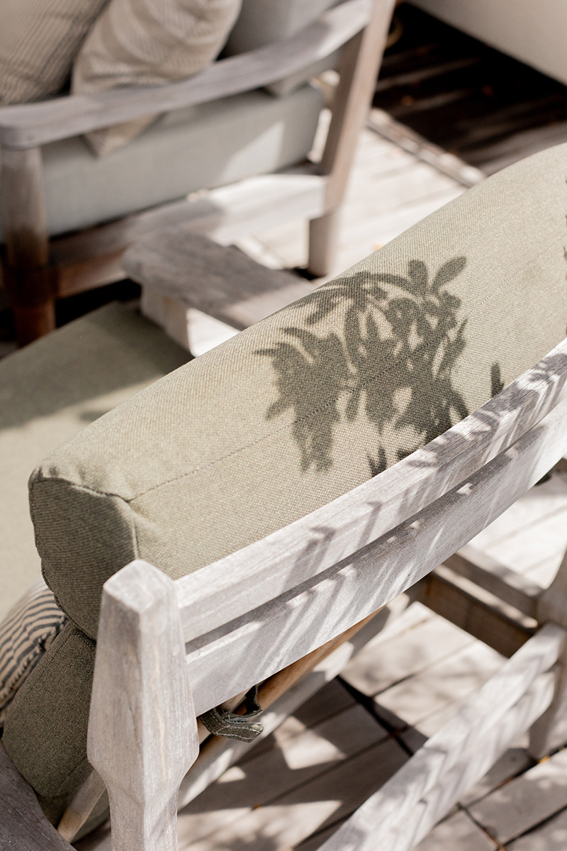 Close-up of a light green outdoor chair with a shadow of leaves cast on the backrest, positioned on a wooden deck.
