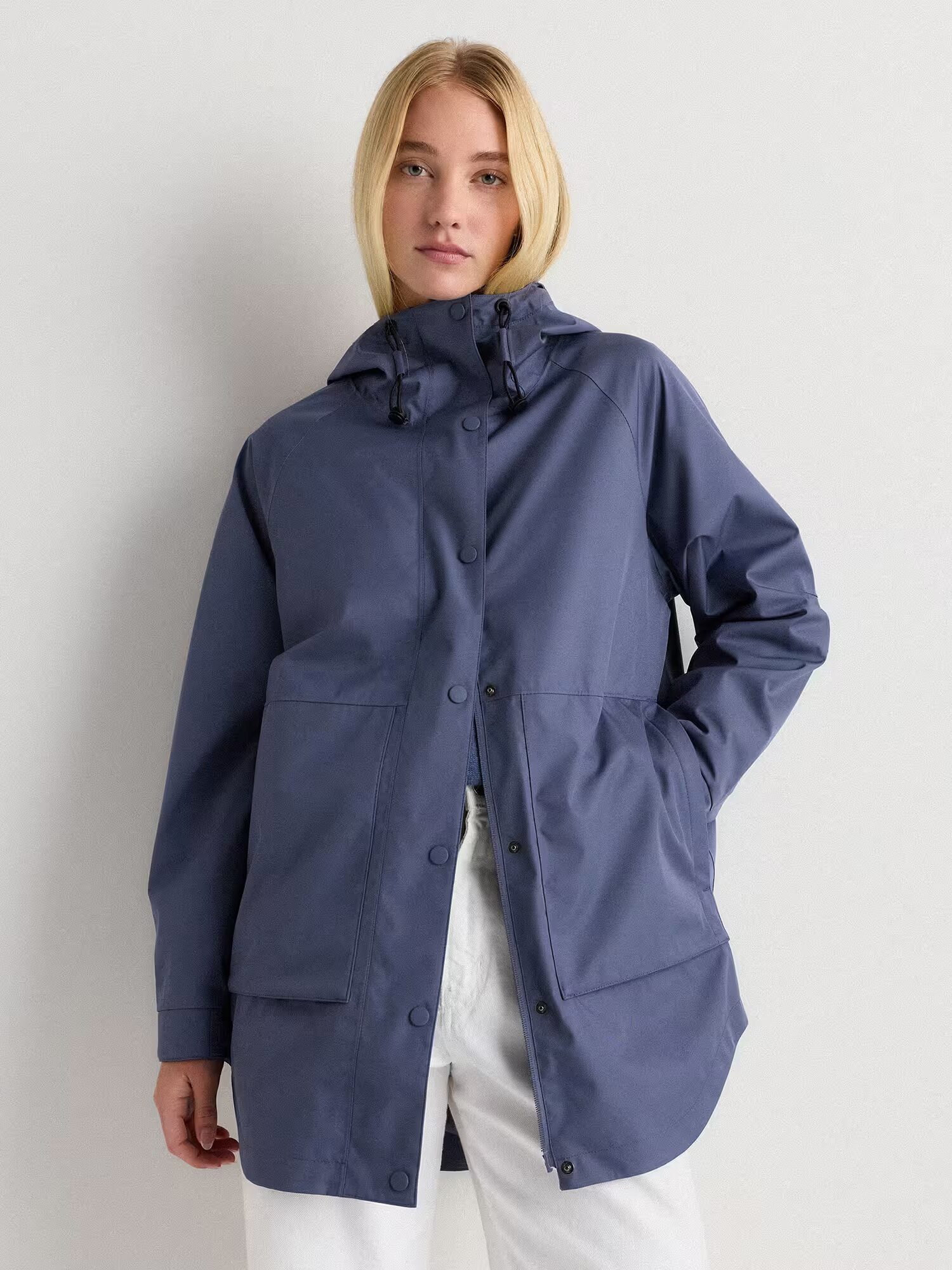 A person wearing a blue waterproof raincoat with large front pockets and white pants stands against a plain light background.