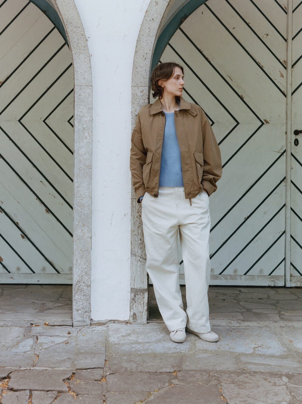 Person wearing a waterproof raincoat, blue sweater, and white pants stands under an archway in front of a wooden, chevron-patterned door, looking to the side.