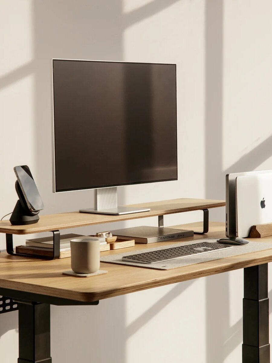 A modern wooden standing desk with a monitor, keyboard, smartphone, mug, closed laptop, and desk organizer in a sunlit room near large windows.