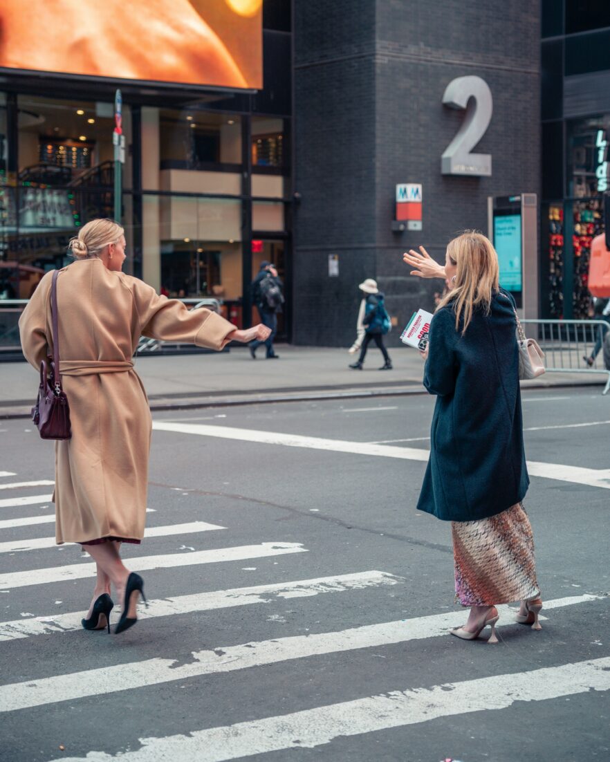Two women wearing coats and heels cross a city street; one raises her arm, possibly signaling for a taxi, while buildings and pedestrians are visible in the background.