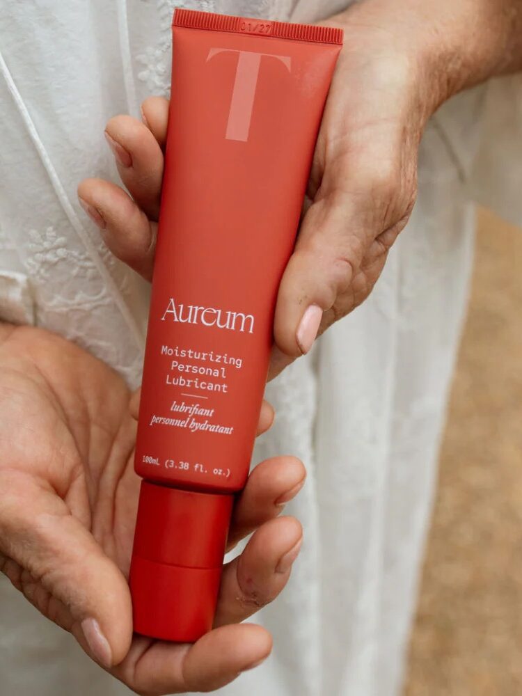 A person in a white garment holds a red tube labeled "Aureum Moisturizing Personal Lubricant" in their hands.