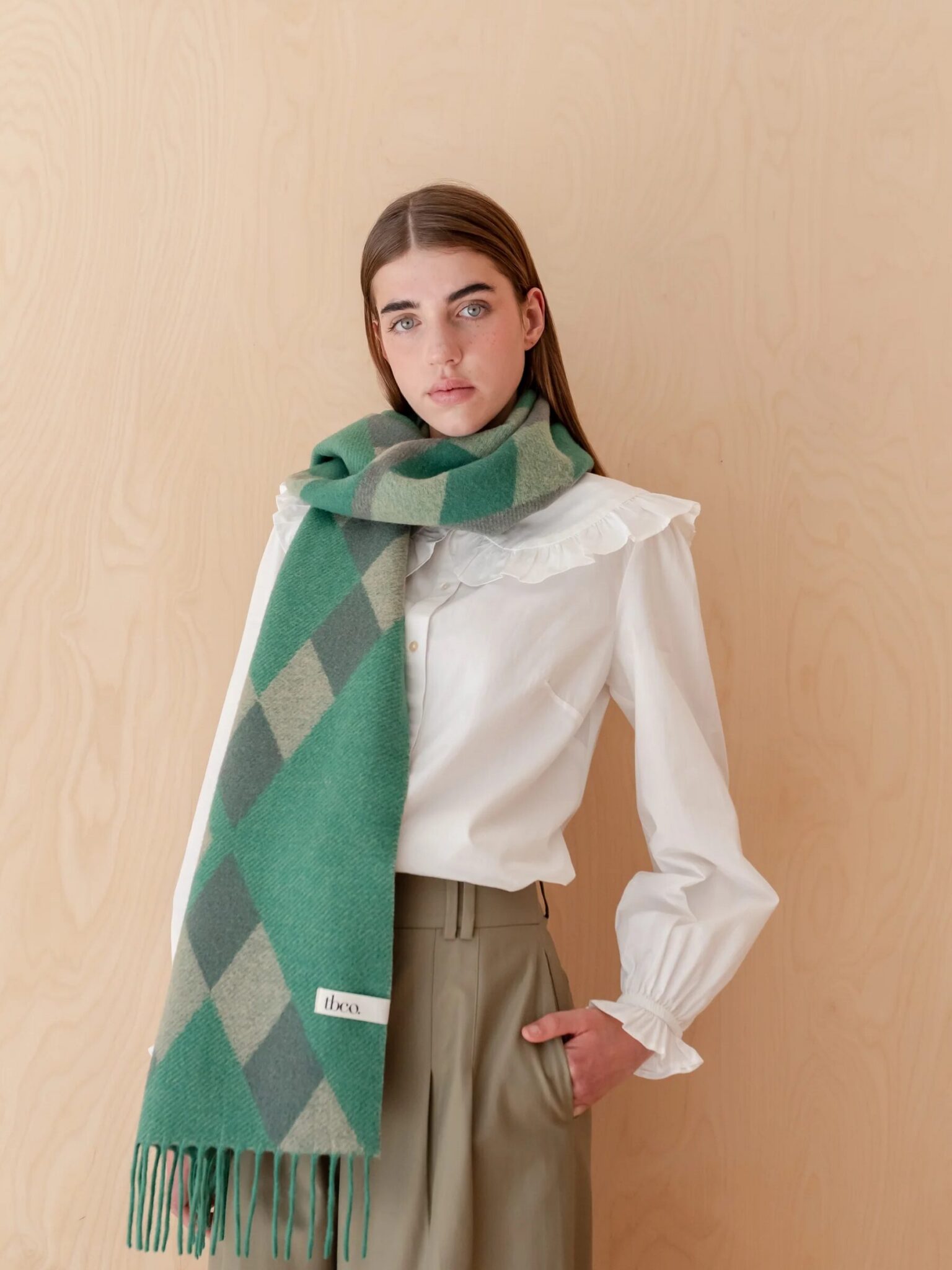 A woman wearing a white blouse and olive trousers stands against a light wood background, with a green argyle scarf draped around her neck.