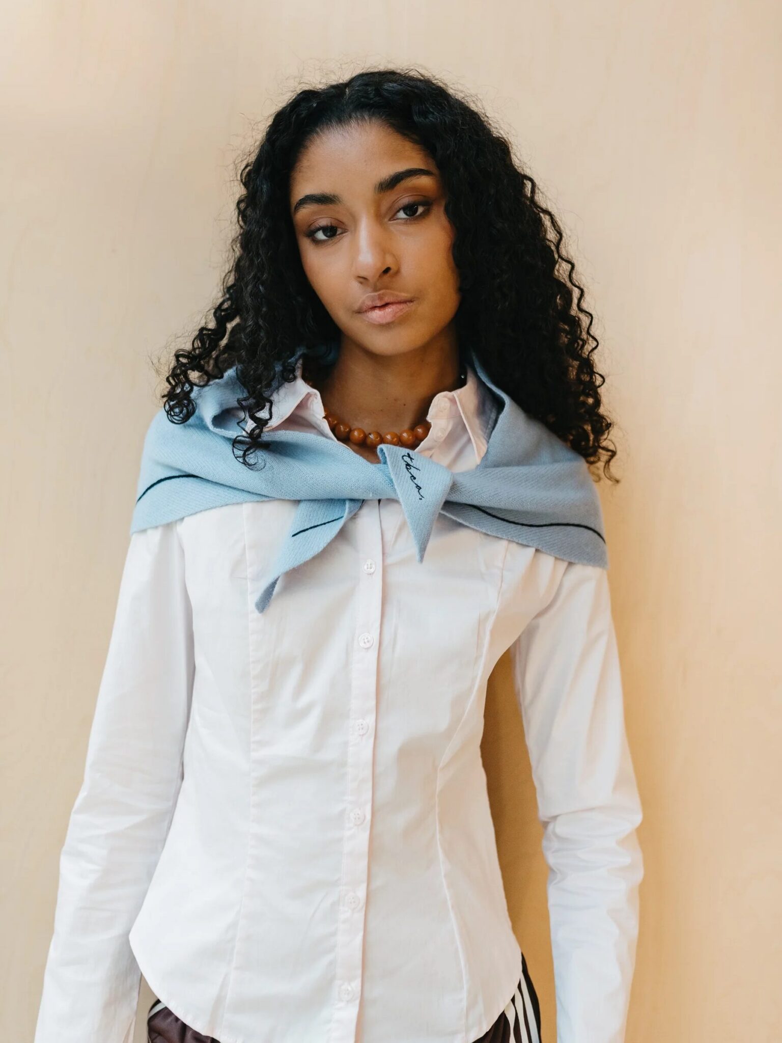 A woman with curly hair wears a white button-up shirt and a light blue sweater draped over her shoulders, standing against a plain beige background.