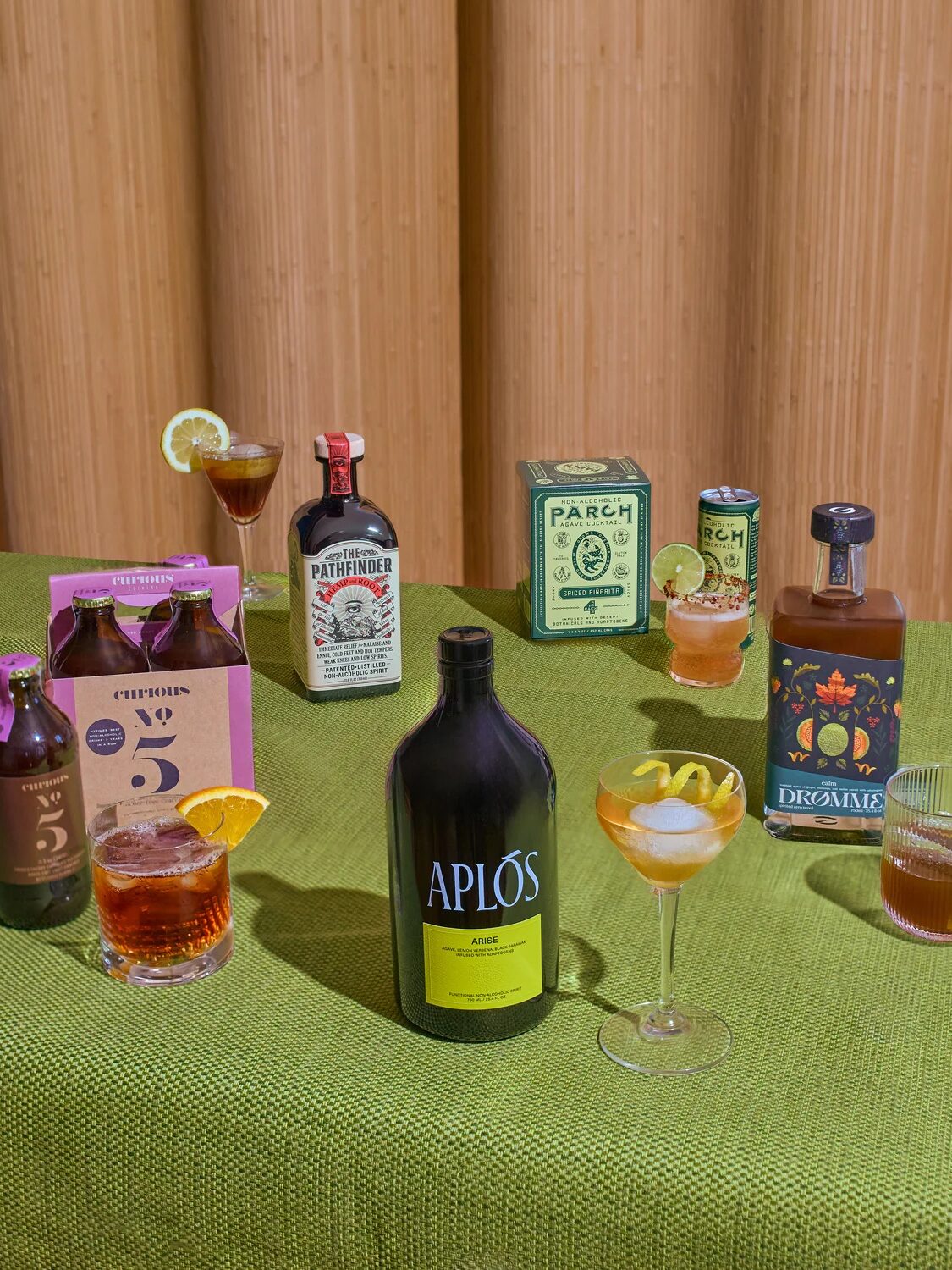 A variety of non-alcoholic spirits and drinks, available for online shopping, are arranged on a green tablecloth, each with a prepared cocktail in a glass beside its respective bottle.