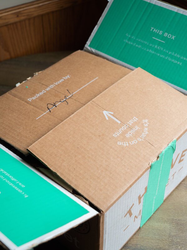 A partially opened cardboard shipping box with green flaps, labeled "Packed with love by Angel," and an arrow pointing to "This side up.