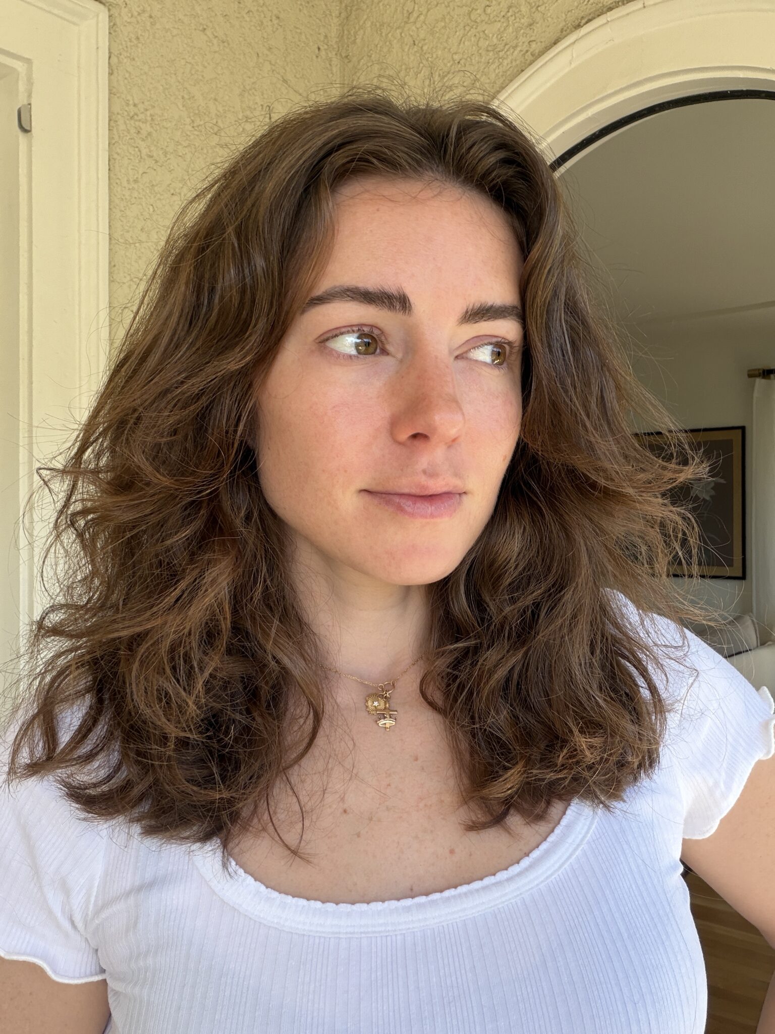 A woman with wavy brown hair and a white top stands indoors, looking slightly to the side with a neutral expression.