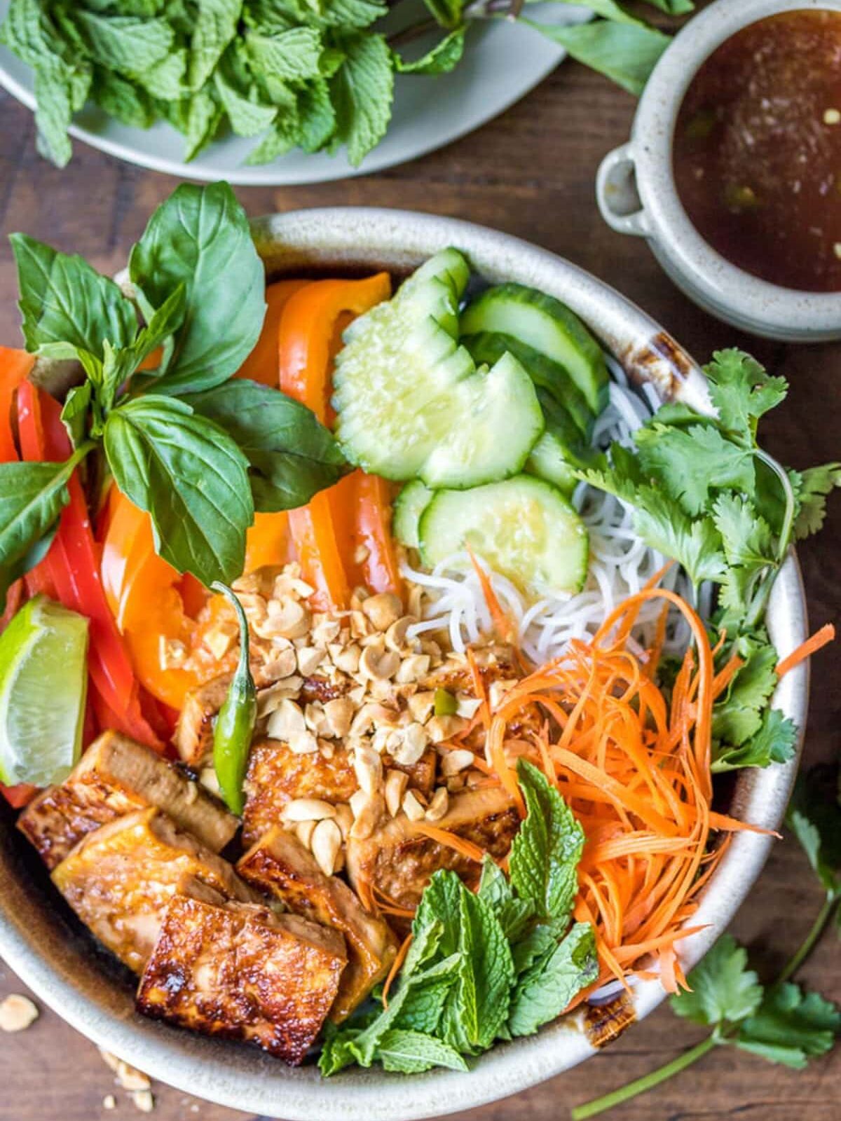 A bowl of Vietnamese vermicelli noodles with grilled tofu, sliced bell peppers, cucumber, carrots, mint, basil, cilantro, lime wedge, and chopped peanuts, with sauce on the side.