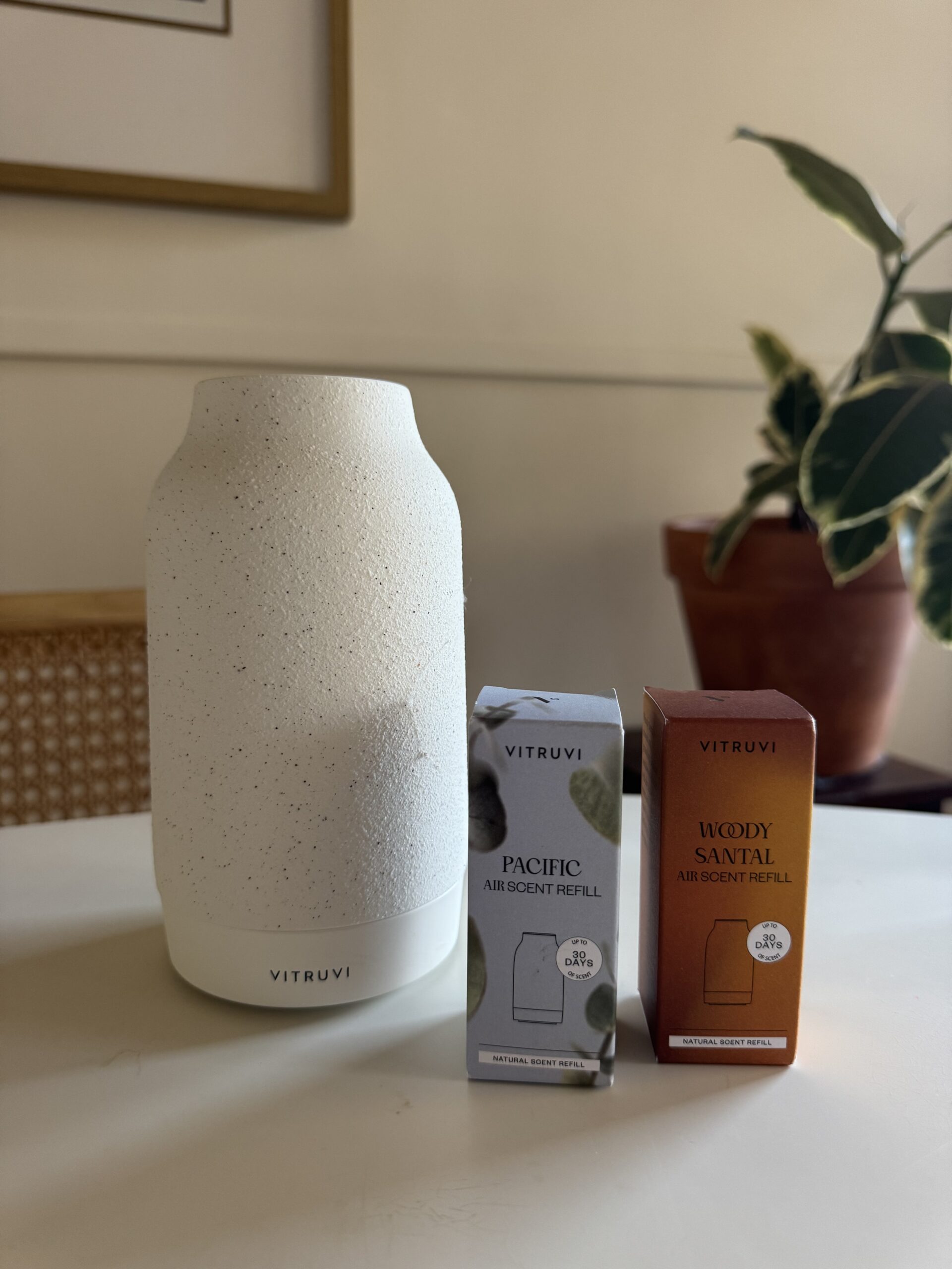 A Vitruvi diffuser sits on a table next to two boxed air scent refills labeled Pacific and Woody Santal. A potted plant and framed art are in the background.