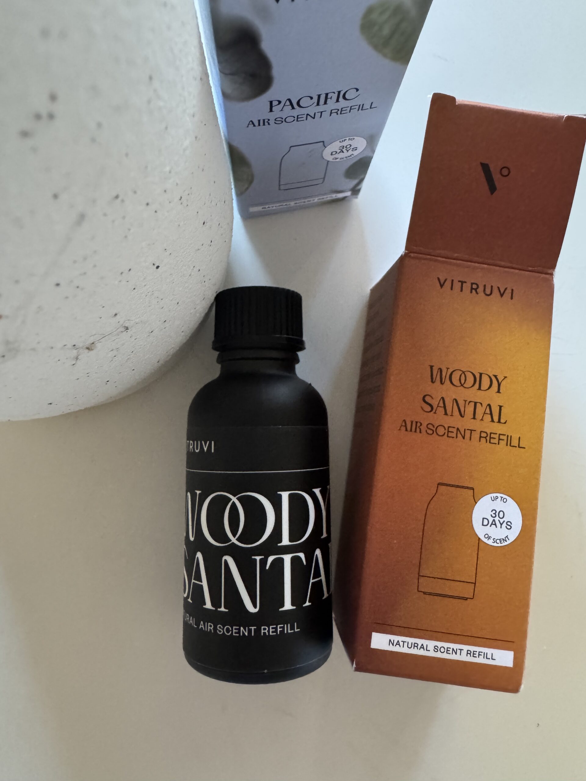 A black bottle labeled “Woody Santal Air Scent Refill” is shown next to its box and a “Pacific Air Scent Refill” box.