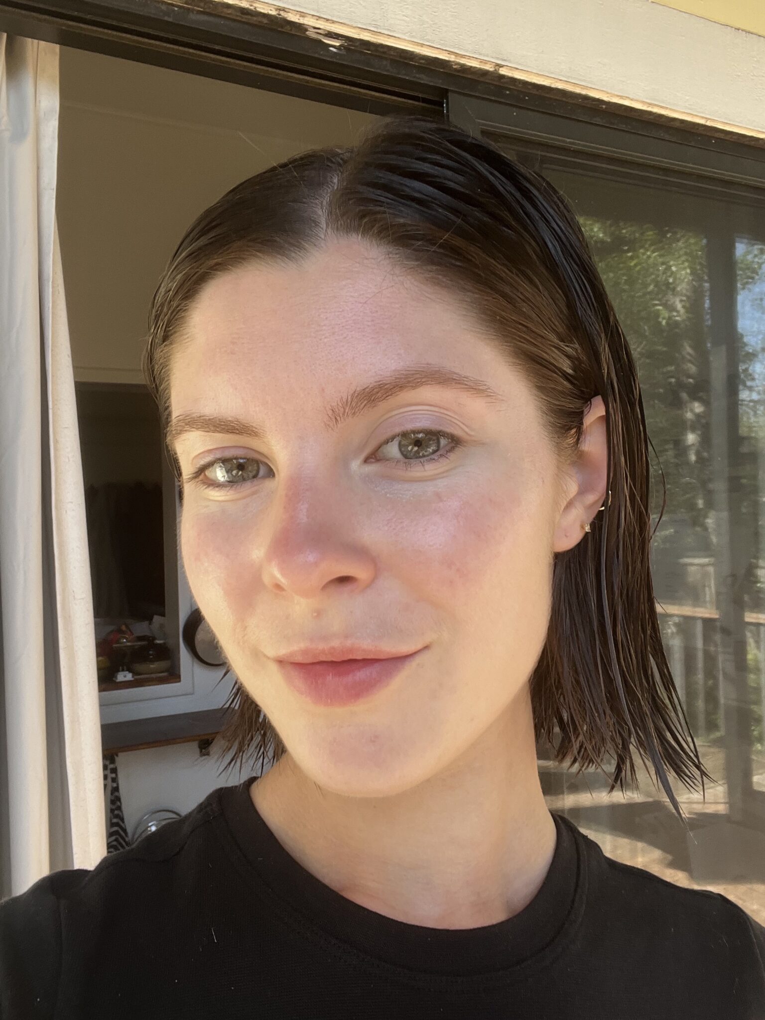 A person with short, wet, dark hair and light skin is standing indoors near a sliding glass door, looking at the camera with a neutral expression.