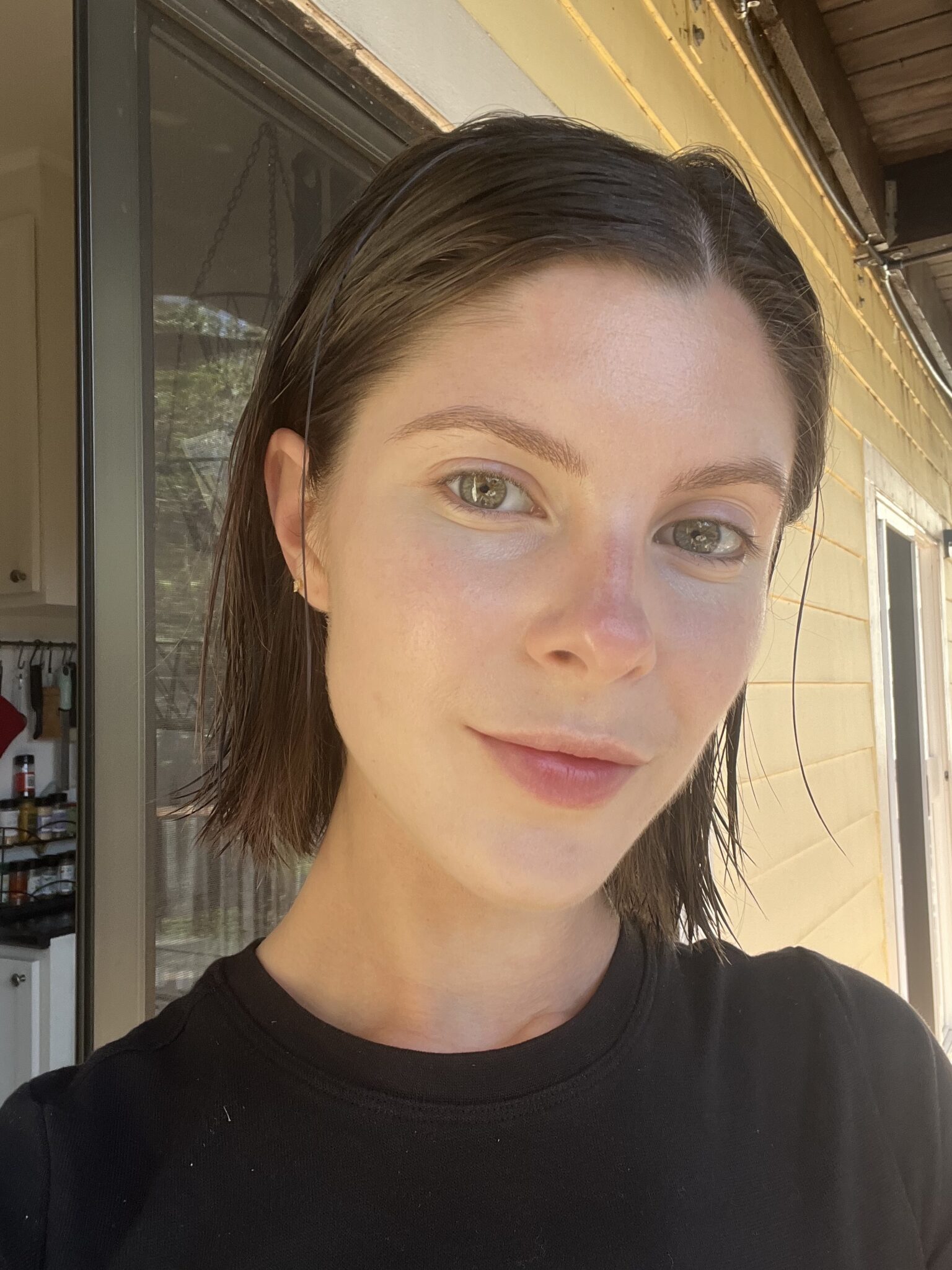 A woman with short brown hair and light skin stands outside near a yellow wall, wearing a black shirt and looking at the camera in natural light.
