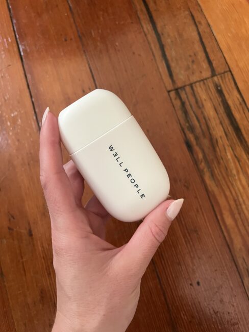 A hand holding a white WELL PEOPLE container against a wooden floor background.