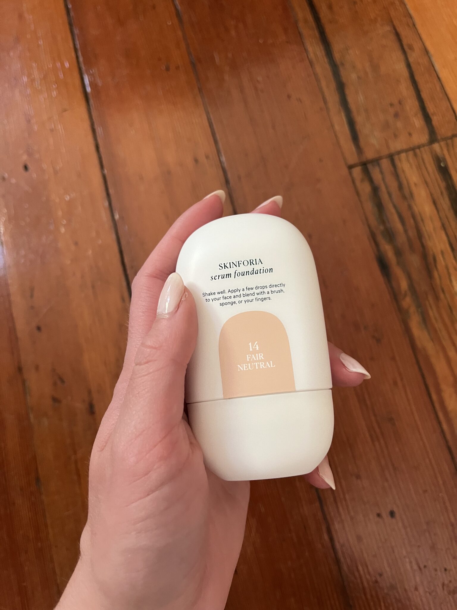 A hand holding a bottle of Skinforia serum foundation in shade 1.4 Fair Neutral, with a wooden floor in the background.