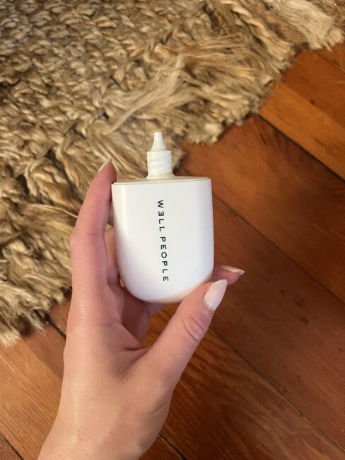A hand holding a white Well People bottle with a nozzle, positioned over a wooden floor and a textured beige rug.