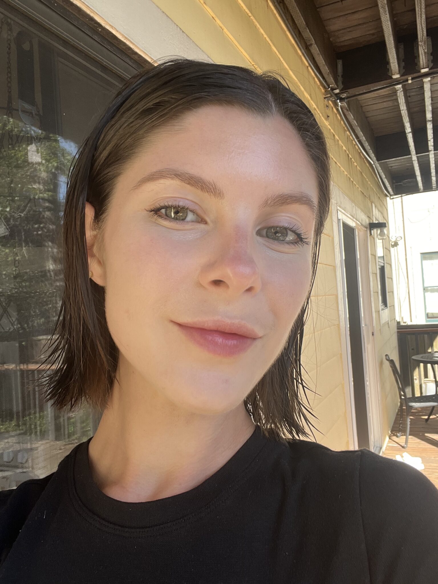 A woman with short brown hair and a black shirt takes a selfie outside near a yellow building on a sunny day.