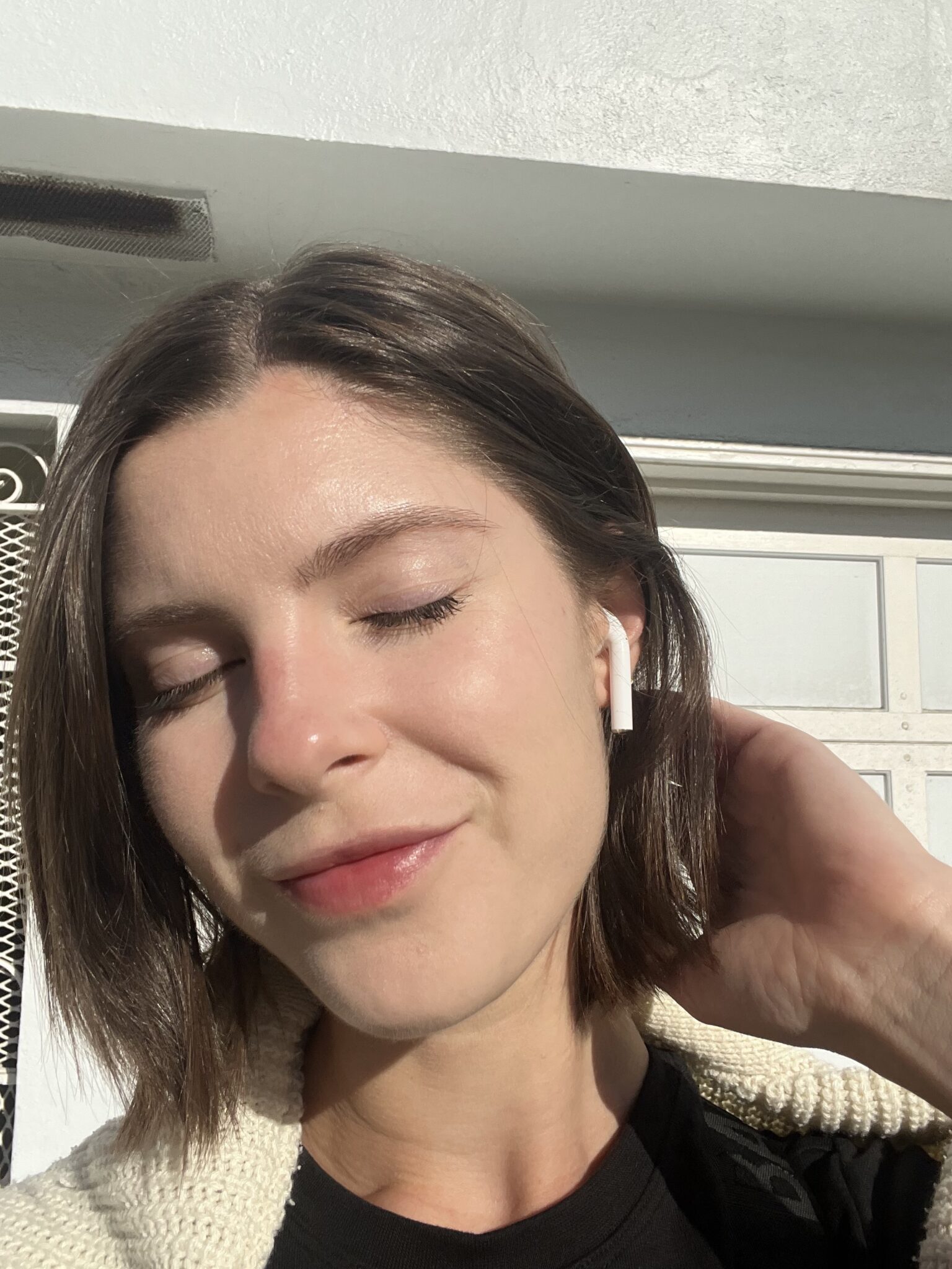 Woman with short brown hair and white earbuds stands outside with eyes closed, smiling slightly in sunlight, hand touching back of her head.