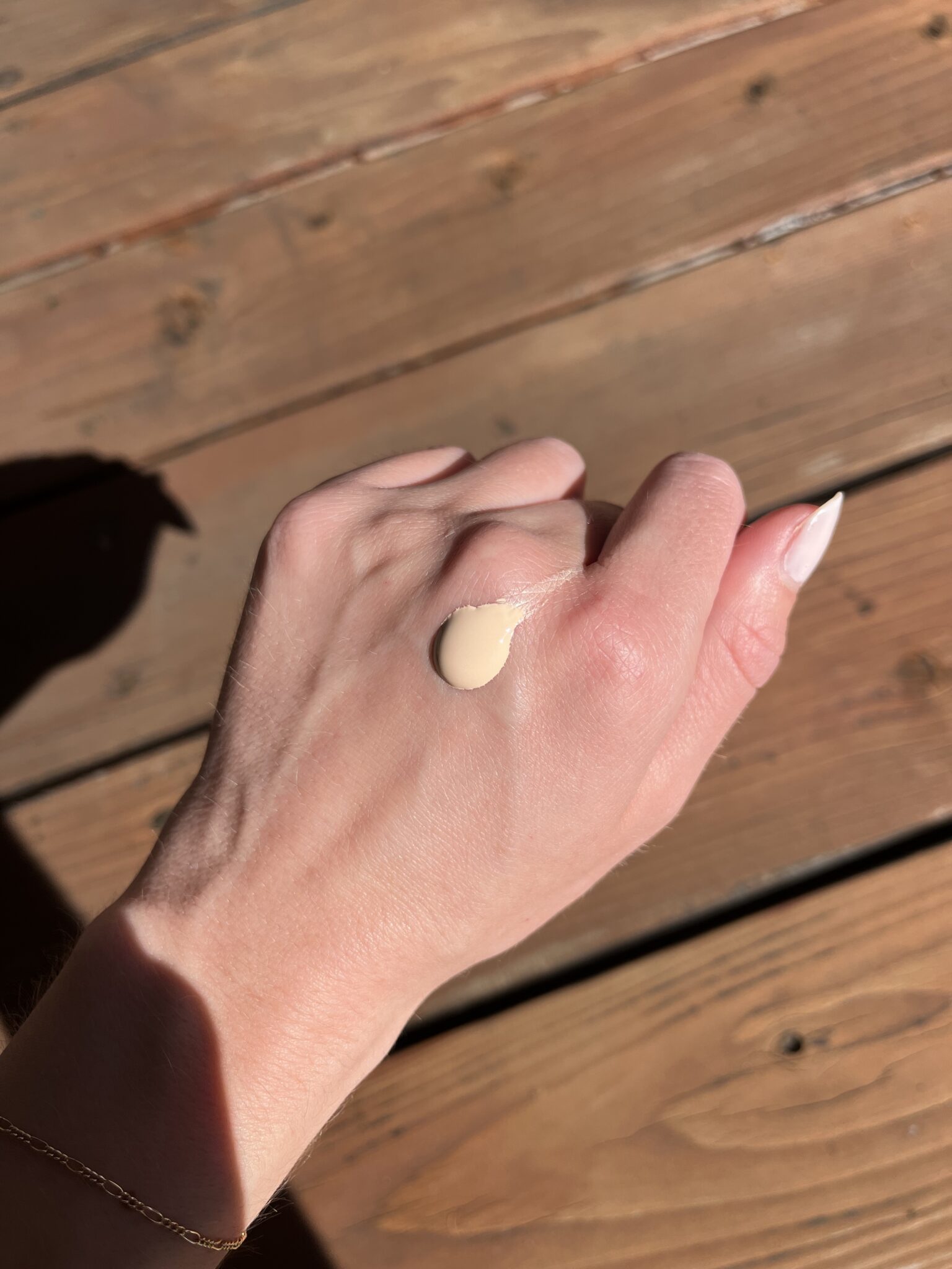 A hand with a drop of beige liquid foundation on the back, shown against a wooden surface in bright sunlight.
