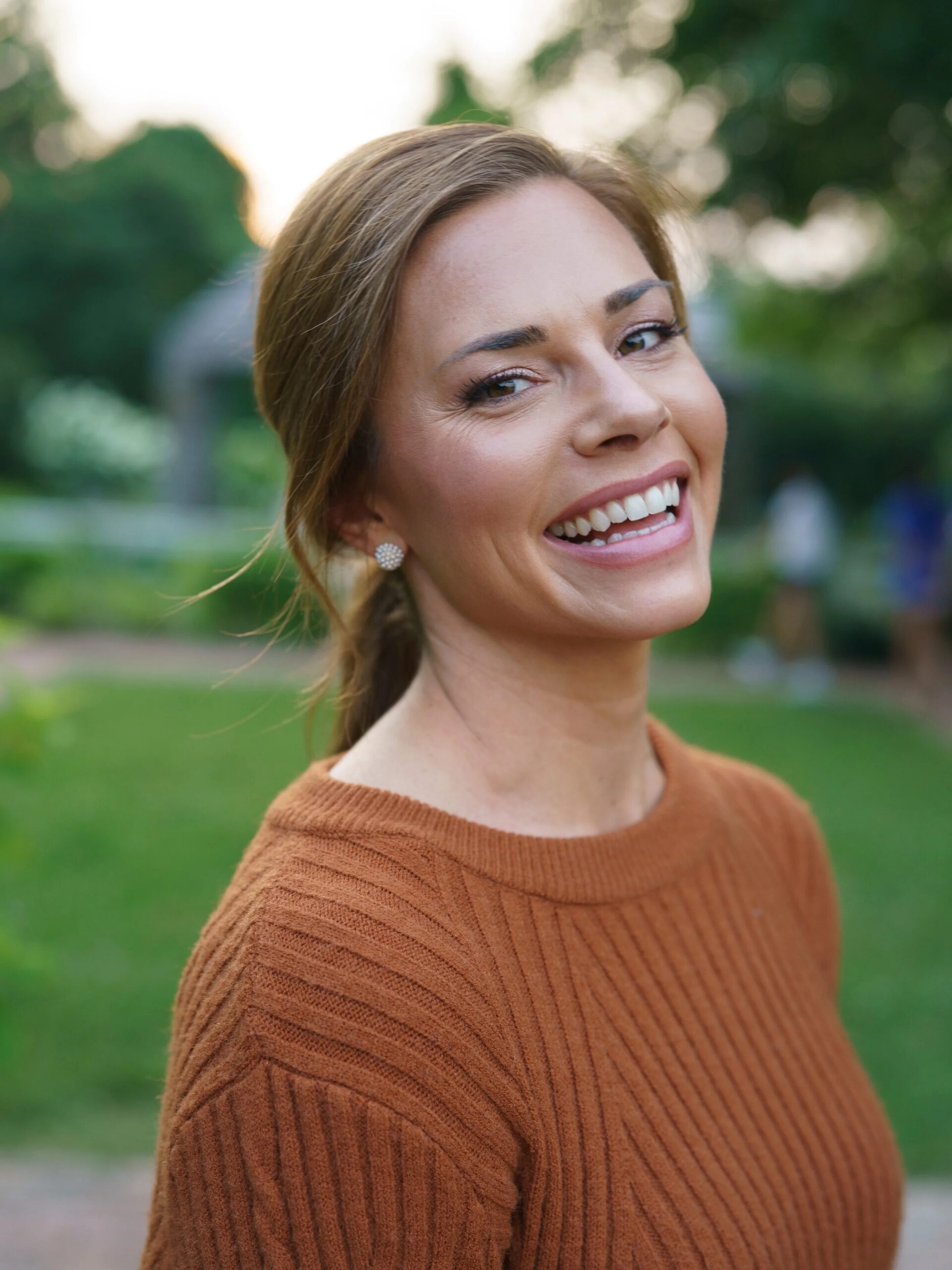 A woman with light brown hair wearing a rust-colored sweater smiles outdoors, with greenery and blurred people in the background.