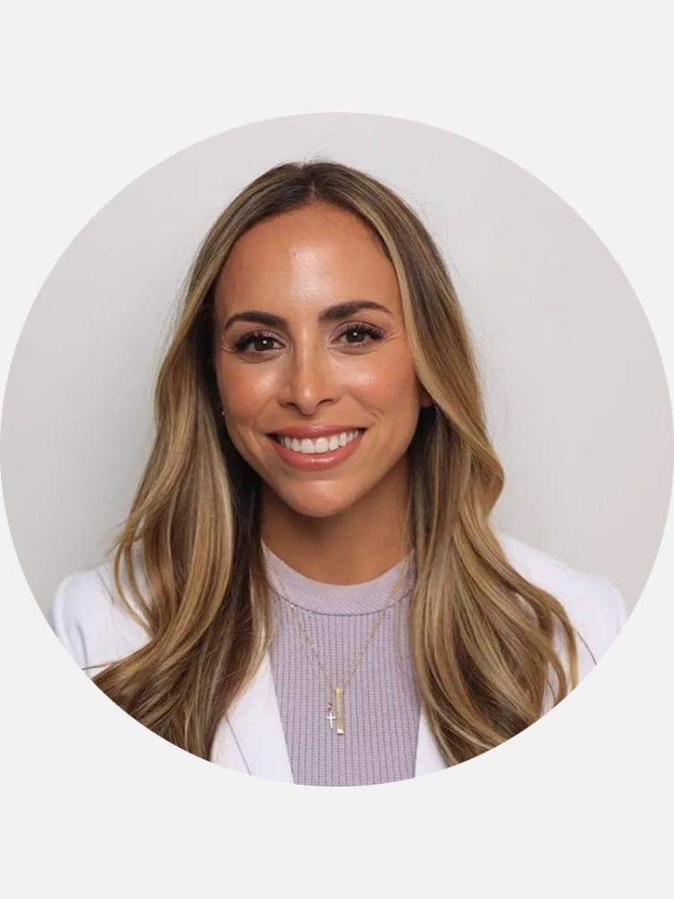 Woman with long, light brown hair wearing a white blazer and lavender top, smiling at the camera against a plain, light background.
