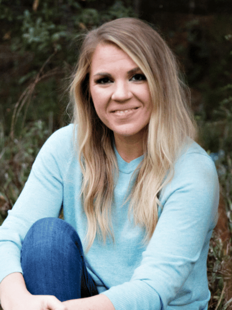 A woman with long blonde hair wearing a light blue sweater and jeans sits outdoors, smiling at the camera with greenery in the background.