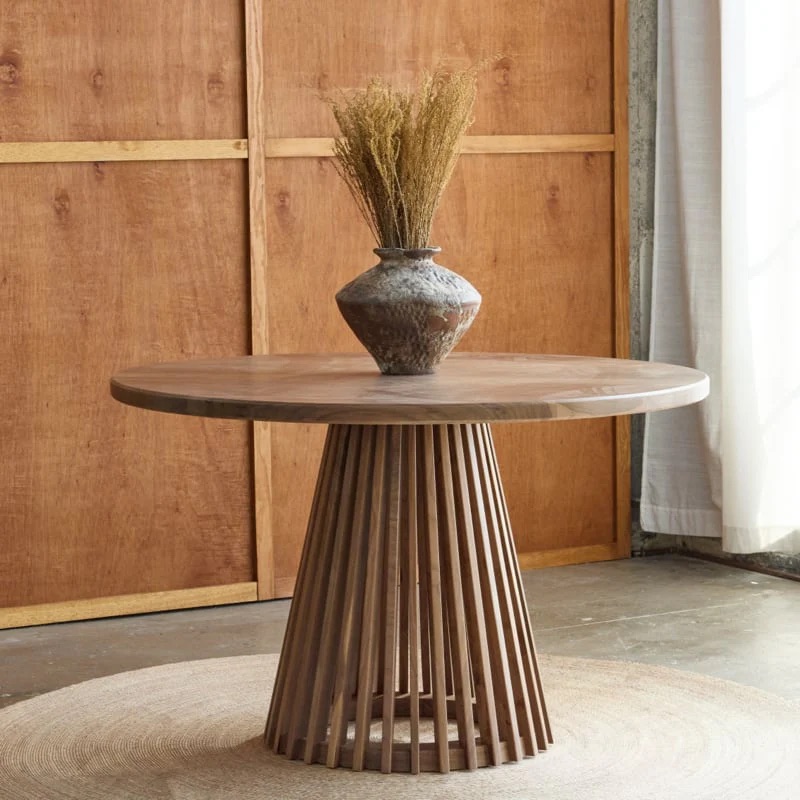 Round wooden table with slatted base, topped with a ceramic vase holding dried grasses, placed on a woven rug in a room with wood-paneled walls.