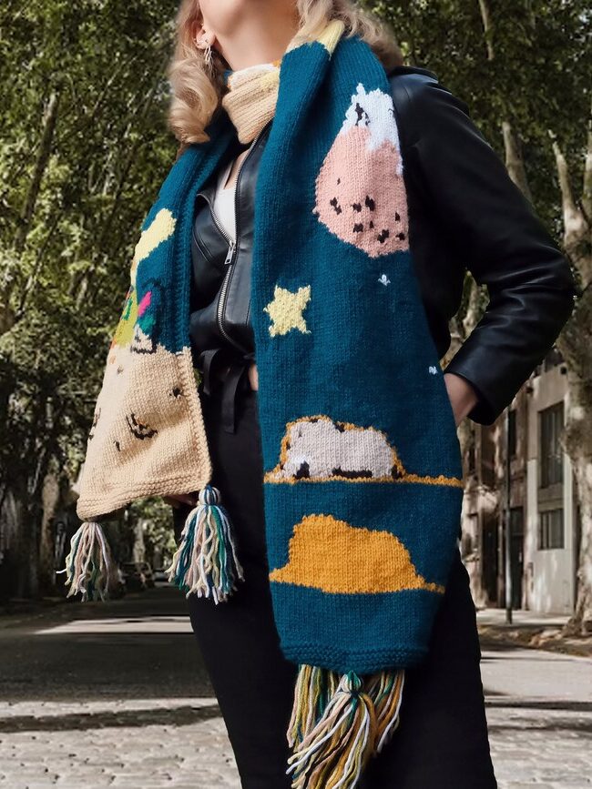 A person stands outdoors wearing a black outfit and a long, colorful knitted scarf decorated with stars, a mountain, and a sheep. Trees and buildings line the background.