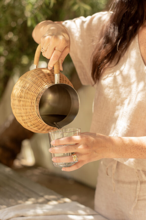 A person in light-colored clothing pours water from a wicker-wrapped pitcher into a clear glass outdoors.