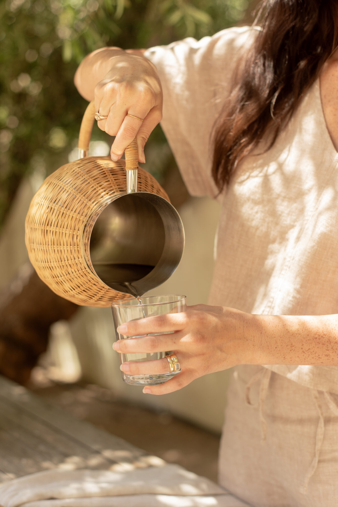 A person in light-colored clothing pours water from a wicker-wrapped pitcher into a clear glass outdoors.