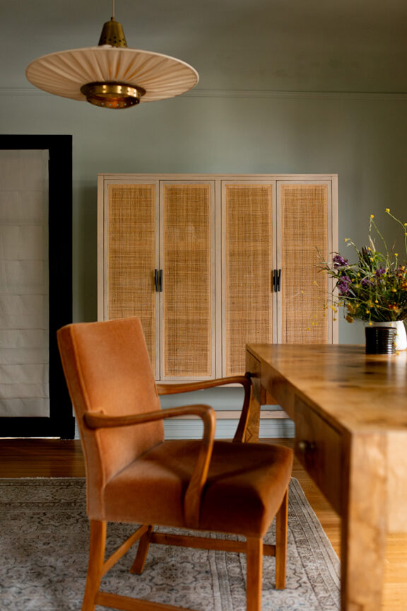A mid-century style office with a wood desk, rust-colored armchair, rattan cabinet, pendant light, and a floral arrangement on the table.
