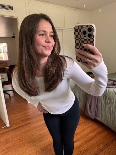 Woman with long brown hair wearing a white top and black pants takes a mirror selfie in a bedroom with wooden floors.
