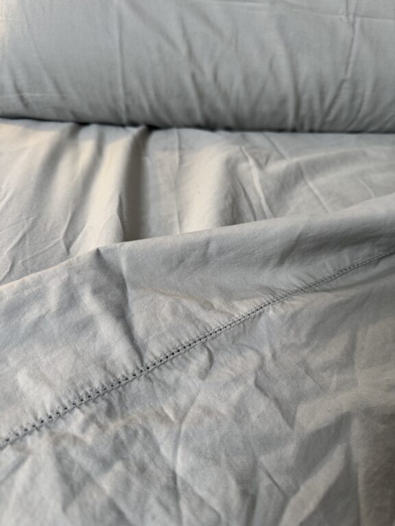 Close-up of light gray, slightly wrinkled bed sheets with visible seam and stitching detail.