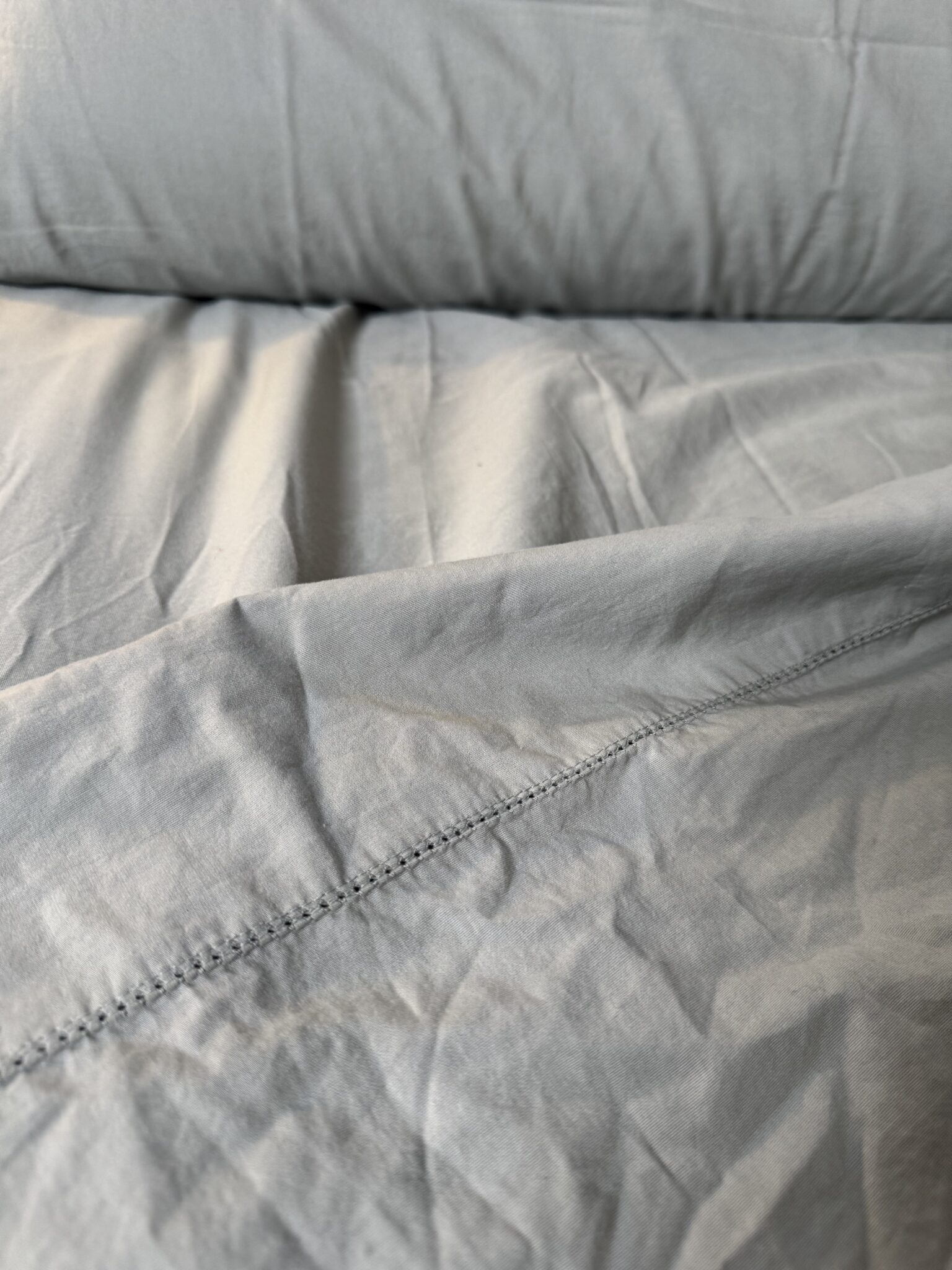 Close-up of light gray, slightly wrinkled bed sheets with visible seam and stitching detail.