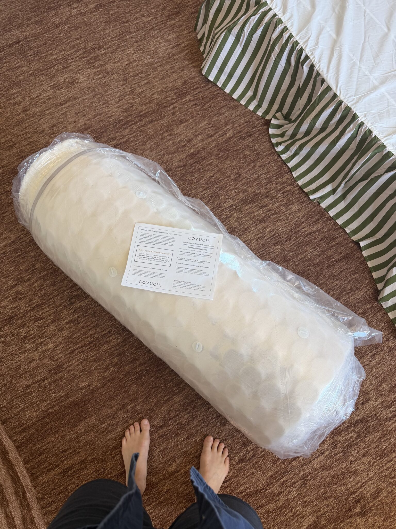 A rolled mattress in plastic packaging with an instruction sheet on top is on a brown carpet next to a bed; bare feet are visible at the bottom of the image.