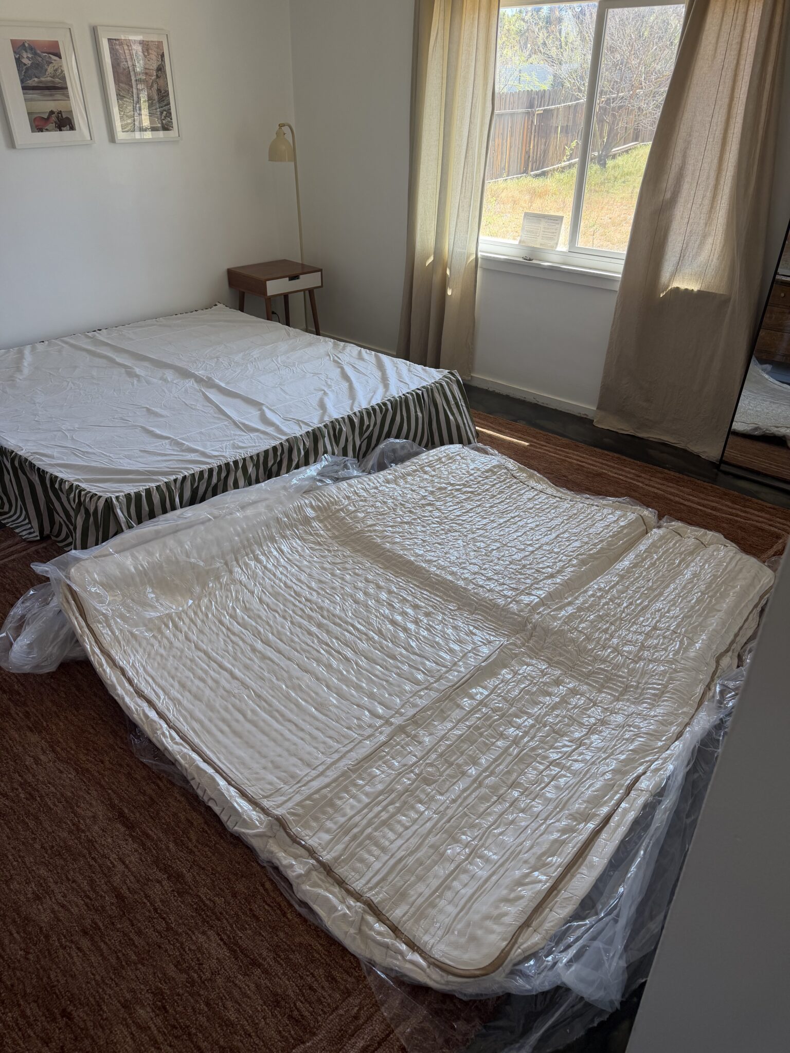 Two mattresses, one on a bed frame and the other in plastic packaging on the floor, are in a bedroom with a window, brown carpet, and framed pictures on the wall.