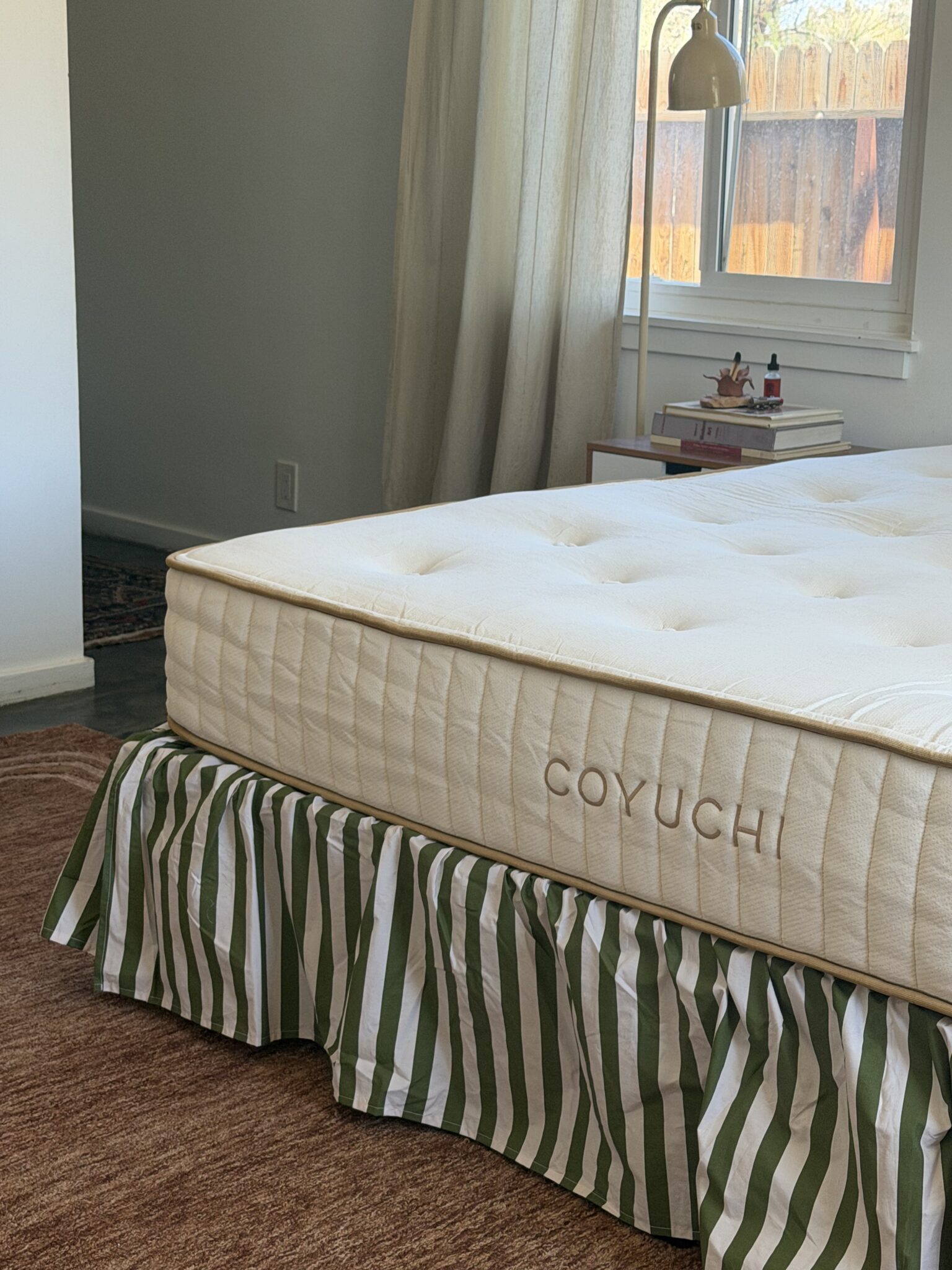 A neatly made bed with a COYUCHI-branded mattress and a green-and-white striped bed skirt in a sunlit bedroom.