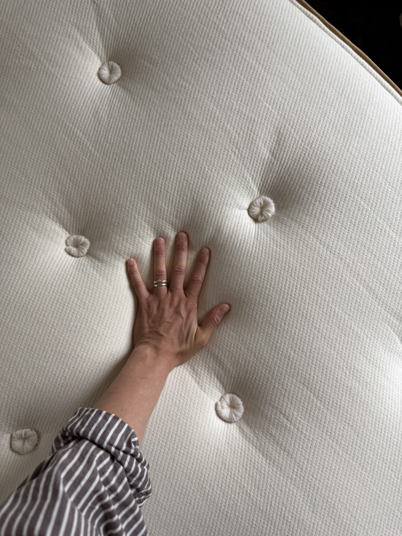A hand presses down on a tufted, off-white mattress surface. The person is wearing a striped sleeve and a ring on their ring finger.