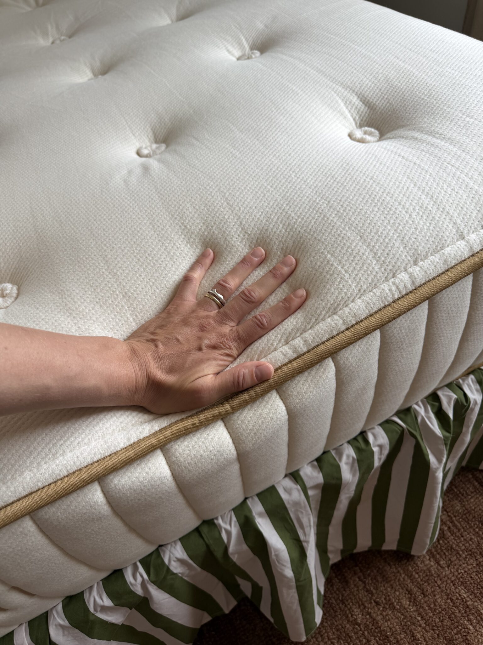 A hand presses down on a white mattress with button tufting and a tan trim, showing its firmness; a green and white striped bed skirt is visible below.