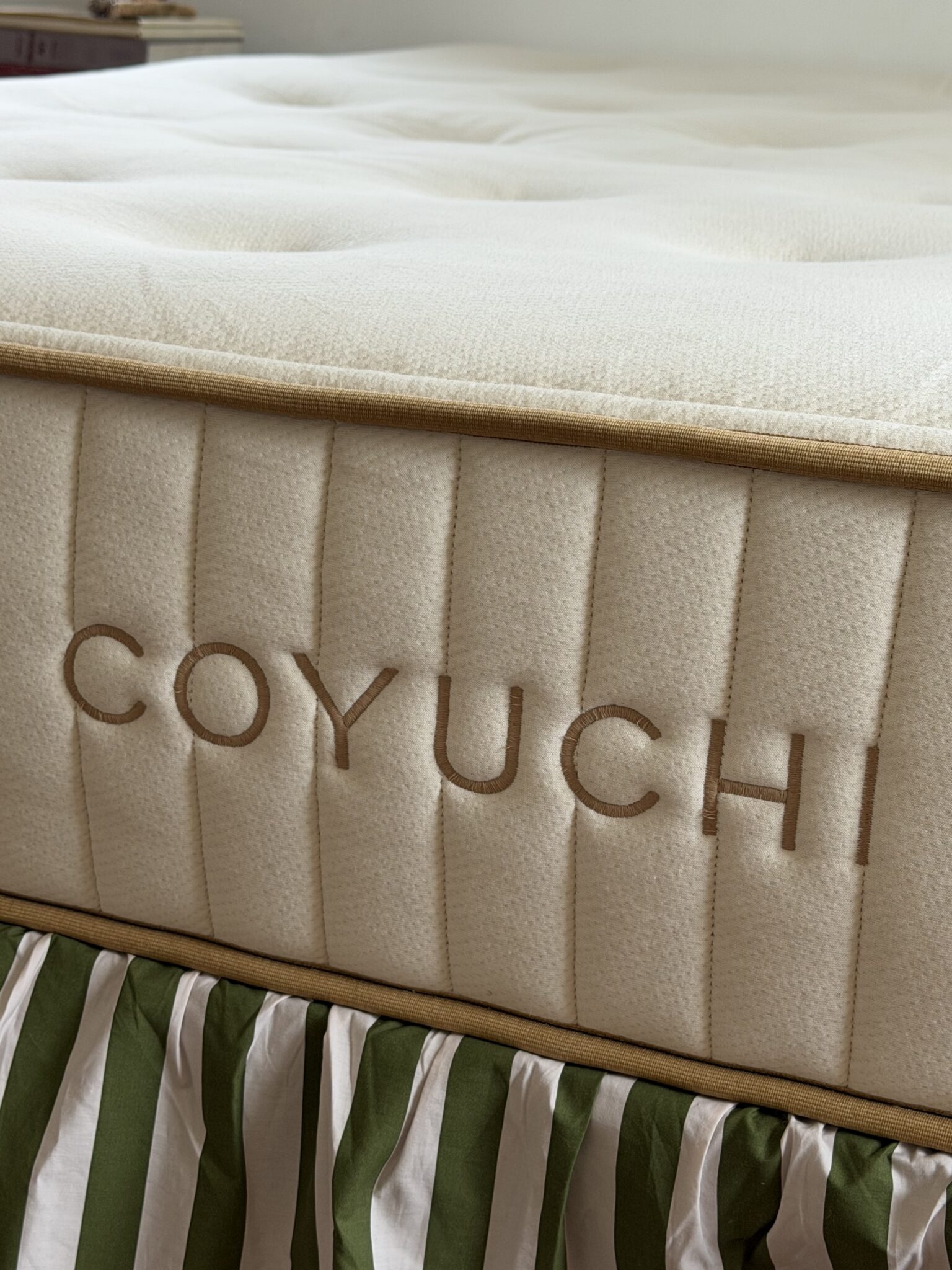 Close-up of a mattress with the brand name "COYUCHI" stitched on the side, resting on a green and white striped bed skirt.