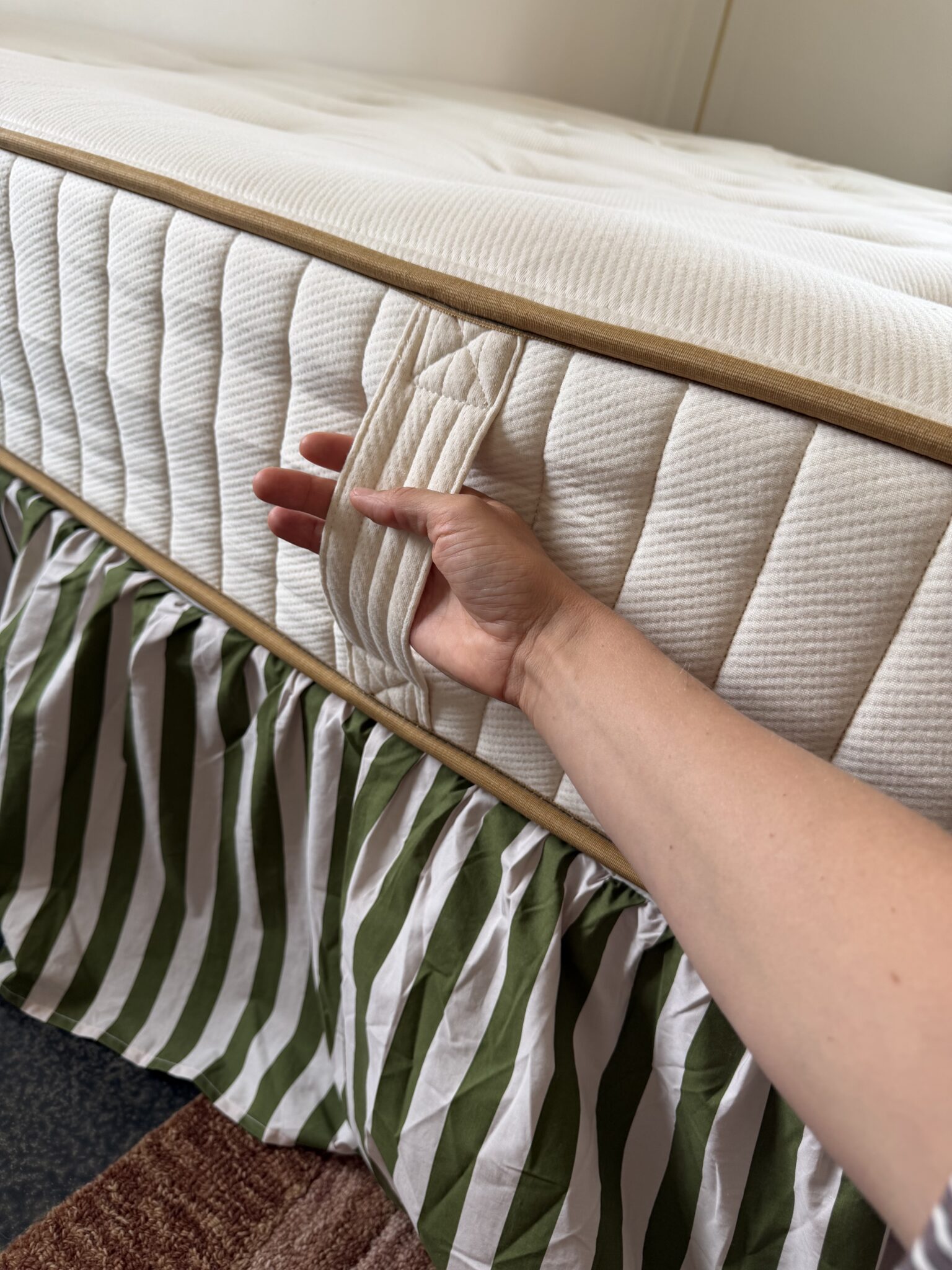 A hand grips the side handle of a mattress with a white and beige cover; a green and white striped bed skirt is partly visible underneath.