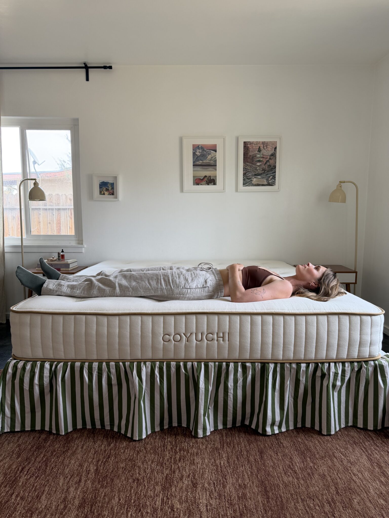 A woman lies on her back on a Coyuchi mattress in a bedroom with white walls, framed art, and a green-striped bed skirt.
