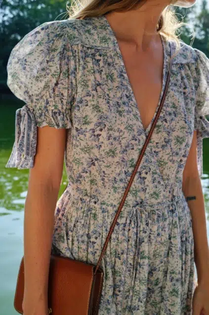 A person wearing a floral dress with puff sleeves and carrying a brown shoulder bag stands near a body of water.