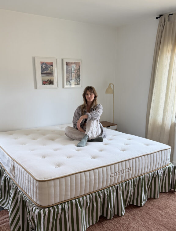 A person in pajamas sits cross-legged on a large Coyuchi mattress with a striped bed skirt in a simply decorated bedroom.
