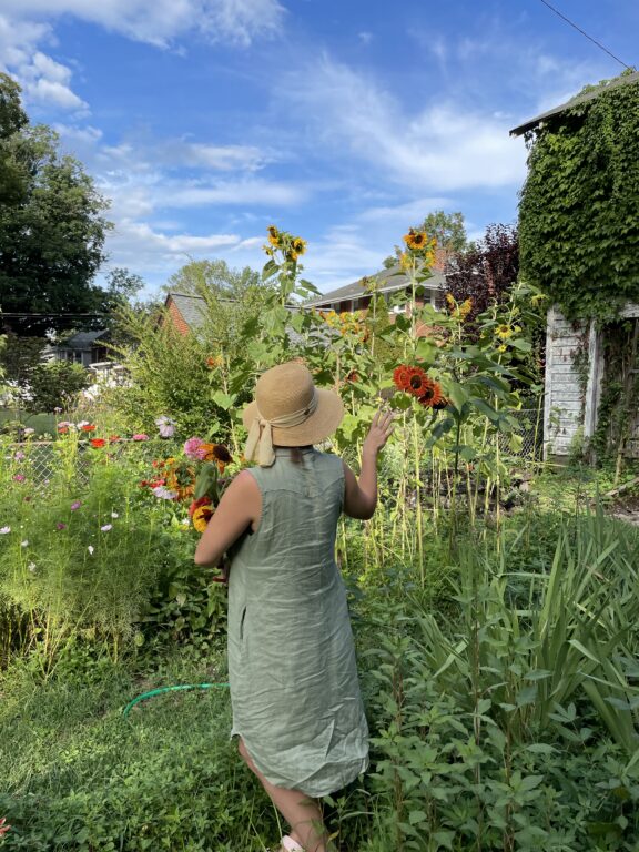 A person in a light green dress and sun hat stands in a garden, holding flowers and facing tall sunflowers under a partly cloudy sky.